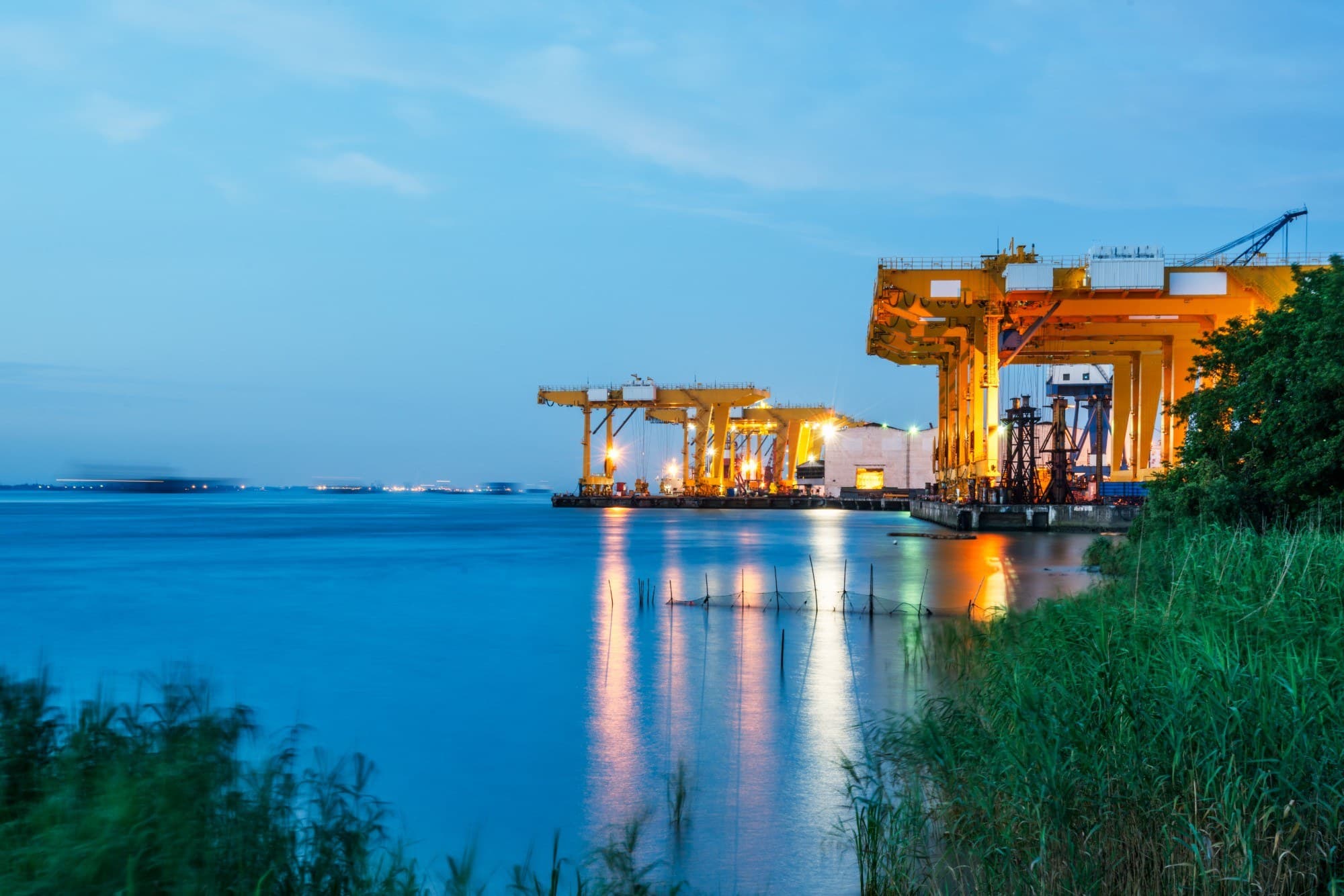 Industrial gantry cranes lit at dusk over a waterfront engineering yard